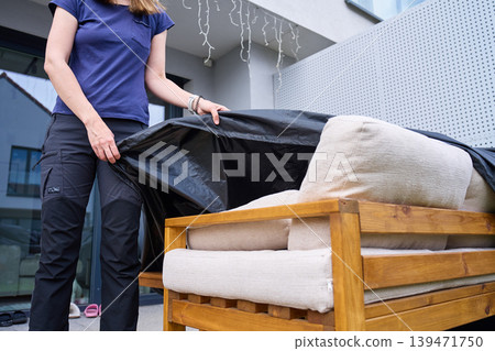 Woman places black protective cover over wooden patio sofa with beige cushions 139471750