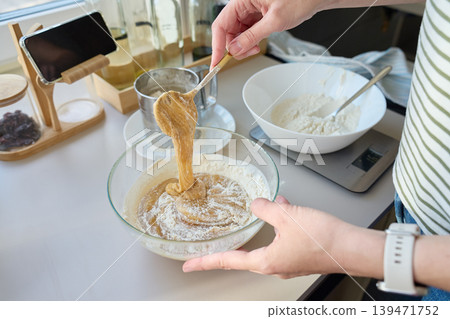 Woman lifts sticky batter with spoon over glass bowl with flour on kitchen counter Woman lifts sticky batter with spoon over glass bowl with flour on kitchen counter 139471752