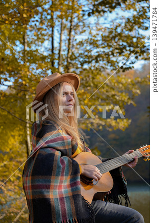 Beautiful blonde girl playing acoustic guitar and singing relaxing enjoying outdoors on sunny autumn day. Hippie woman in boho style learning and practicing playing musical instrument. Fall leaves Beautiful blonde girl playing acoustic guitar and singing relaxing enjoying outdoors on sunny autumn day. Hippie woman in boho style learning and practicing playing musical instrument. Fall leaves 139471784