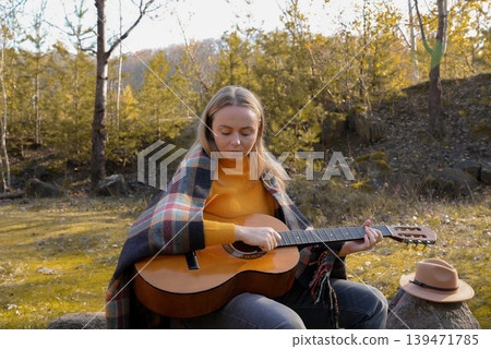 Beautiful blonde girl playing acoustic guitar and singing relaxing enjoying outdoors on sunny autumn day. Hippie woman in boho style learning and practicing playing musical instrument. Fall leaves Beautiful blonde girl playing acoustic guitar and singing relaxing enjoying outdoors on sunny autumn day. Hippie woman in boho style learning and practicing playing musical instrument. Fall leaves 139471785