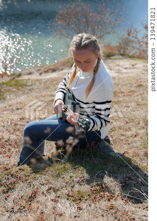 Portrait of smiling caucasian girl plays ukulele in autumnal park. Young woman plays guitar musical instrument outside in nature fall time. Audio music healing Portrait of smiling caucasian girl plays ukulele in autumnal park. Young woman plays guitar musical instrument outside in nature fall time. Audio music healing 139471821