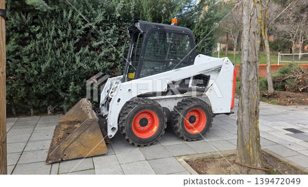 Side view skid steer loader with bucket and orange wheels parked near trees for road maintenance and construction background use 139472049