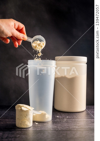 Protein drink. Hand pouring protein powder in a white shaker. Plastic tube on dark background. Body building, muscle growth and food supplement concept Protein drink. Hand pouring protein powder in a white shaker. Plastic tube on dark background. Body building, muscle growth and food supplement concept 139472057