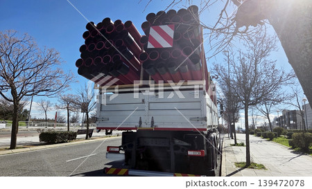 Rear view of a truck carrying stacked red pipes on city road for road repair and infrastructure transport background Rear view of a truck carrying stacked red pipes on city road for road repair and infrastructure transport background 139472078