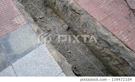 Close up urban sidewalk trench for pipe laying and road repair behind safety barriers for construction documentation and maintenance background Close up urban sidewalk trench for pipe laying and road repair behind safety barriers for construction documentation and maintenance background 139472184