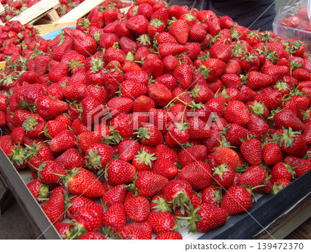 Large, abundant heap of fresh, ripe strawberries fills a market display tray. The vibrant red berries, many with green leafy hulls still attached, present a juicy and natural texture Large, abundant heap of fresh, ripe strawberries fills a market display tray. The vibrant red berries, many with green leafy hulls still attached, present a juicy and natural texture 139472370