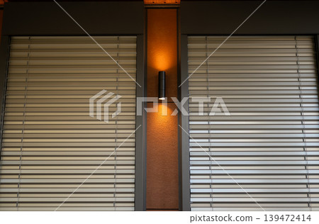 Modern building facade featuring metal external blinds and a warm vertical wall lamp between windows. Modern building facade featuring metal external blinds and a warm vertical wall lamp between windows. 139472414