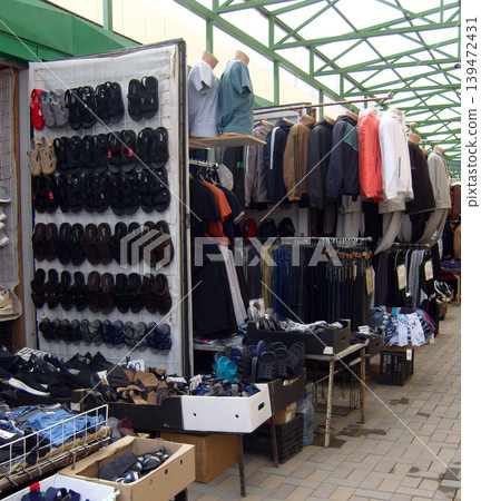 Bustling outdoor market stall displays wide variety of affordable clothing and footwear. Rows of sandals hang on white grid, while racks of jackets, shirts, and trousers sit under a green metal roof Bustling outdoor market stall displays wide variety of affordable clothing and footwear. Rows of sandals hang on white grid, while racks of jackets, shirts, and trousers sit under a green metal roof 139472431