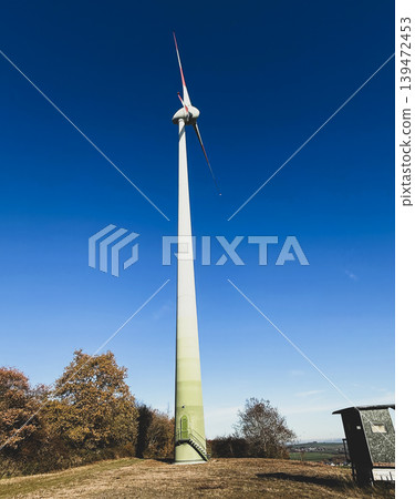 Tall wind turbine rises into deep blue sky above autumn landscape. Clean geometry creates strong vertical perspective Tall wind turbine rises into deep blue sky above autumn landscape. Clean geometry creates strong vertical perspective 139472453
