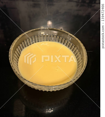 Warm yellow cream in a transparent glass bowl on a dark kitchen surface. Minimal food composition with soft reflections and smooth texture Warm yellow cream in a transparent glass bowl on a dark kitchen surface. Minimal food composition with soft reflections and smooth texture 139472466