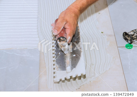 Tiler worker uses trowel to spread adhesive on floor surface during bathroom renovation task at home. Tiler worker uses trowel to spread adhesive on floor surface during bathroom renovation task at home. 139472849