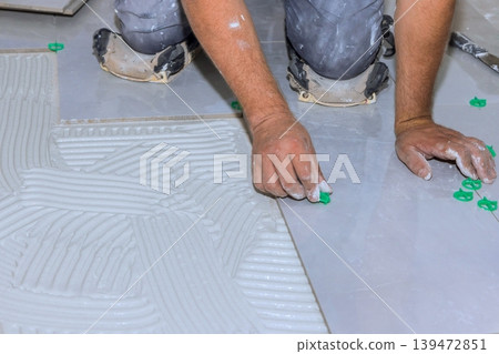 Worker places tiles on floor using cement while adjusting spacers for alignment in home renovation project. Worker places tiles on floor using cement while adjusting spacers for alignment in home renovation project. 139472851