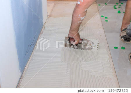 Handy man spreads tile adhesive on floor while working in home interior during renovation stage. Handy man spreads tile adhesive on floor while working in home interior during renovation stage. 139472853