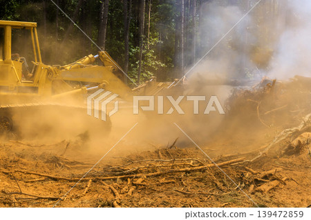 Bulldozer clears fallen trees in forest, kicking up dust debris while smoke rises in air 139472859
