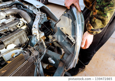Hands work on repairing car headlight parts removed in garage with other vehicles nearby. Hands work on repairing car headlight parts removed in garage with other vehicles nearby. 139472865