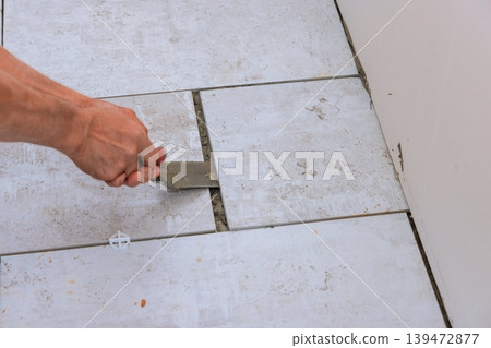 Hand holds trowel while filling gaps between tiles at kitchen worker task in new home setting. Hand holds trowel while filling gaps between tiles at kitchen worker task in new home setting. 139472877