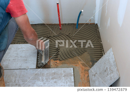 Worker spreads mortar on floor while preparing to install tiles in bathroom during home renovation work. Worker spreads mortar on floor while preparing to install tiles in bathroom during home renovation work. 139472879