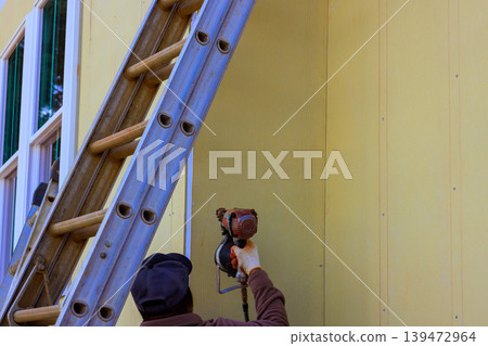 Worker uses pneumatic nail gun to installing siding outside of building while standing on ladder Worker uses pneumatic nail gun to installing siding outside of building while standing on ladder 139472964