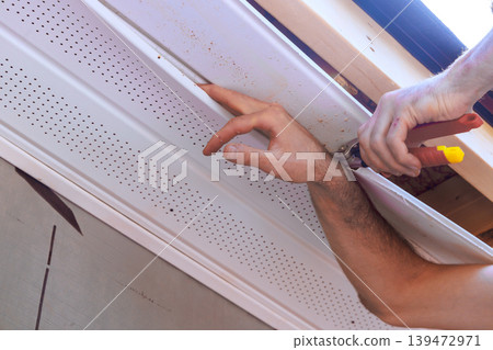 Carpenter uses cutting tools to fix something soffit trim to ceiling standing outdoors Carpenter uses cutting tools to fix something soffit trim to ceiling standing outdoors 139472971