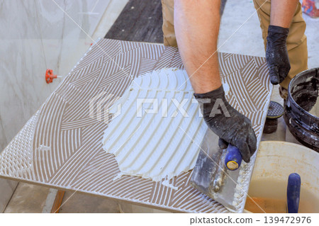 Tiling worker uses trowel to spread adhesive on tile during flooring project in construction area. Tiling worker uses trowel to spread adhesive on tile during flooring project in construction area. 139472976
