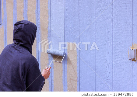 Worker is rolling blue paint on wooden siding wall in home setting during works day. 139472995