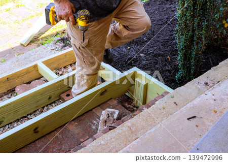 Worker is using drill to secure steps wood pieces while standing on step outside, surrounded by construction materials. 139472996