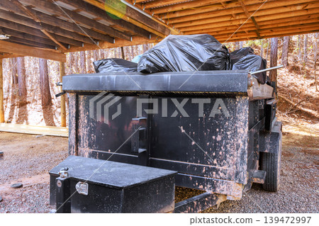 Black dumpster is located under wooden shelter it is filled with garbage bags 139472997