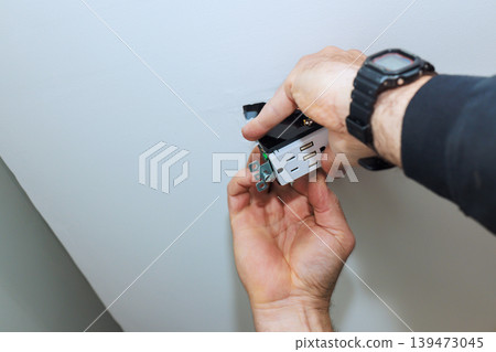 Contractor is working on installing outlet into ceiling while focusing on task in living room setting. Contractor is working on installing outlet into ceiling while focusing on task in living room setting. 139473045