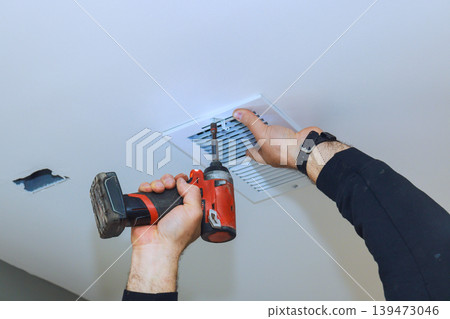Contractor is using power screwdriver to attach vent cover to ceiling in room with hole nearby. Contractor is using power screwdriver to attach vent cover to ceiling in room with hole nearby. 139473046