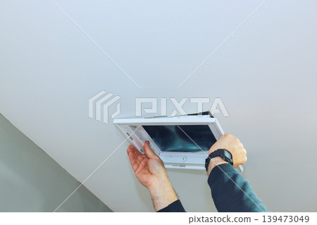 Contractor is taking off an access ceiling vent panel from ceiling while standing in room. Contractor is taking off an access ceiling vent panel from ceiling while standing in room. 139473049