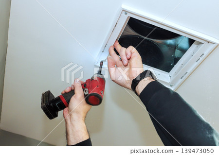 Handy worker uses power drill to secure vent cover to ceiling inside building 139473050
