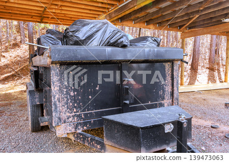 Garbage truck parked under wooden shelter filled with black trash bags in wooded area 139473063