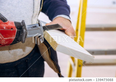 Contractor operates power hand saw on apiece of wood worker stands beside ladder of ongoing construction renovations. 139473088