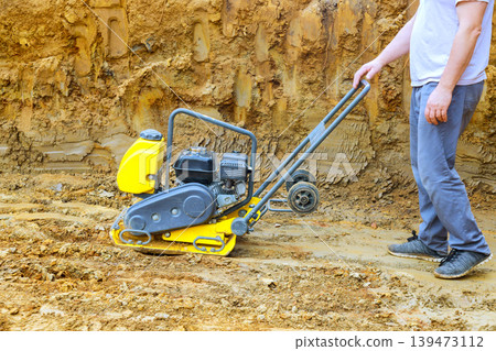 Construction worker pushes plate compactor over loose soil at building site in dust rises as machine flattens ground for paving. 139473112