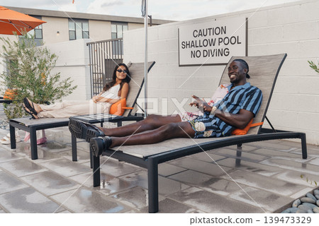 Couple Lounging On Rooftop Pool Deck, Sharing Drinks And Conversation, Orange Cushions And Sunglasses, Modern Tile Flooring, Visible Safety Sign On Wall, Relaxed Summer Mood, Hotel Terrace Couple Lounging On Rooftop Pool Deck, Sharing Drinks And Conversation, Orange Cushions And Sunglasses, Modern Tile Flooring, Visible Safety Sign On Wall, Relaxed Summer Mood, Hotel Terrace 139473329