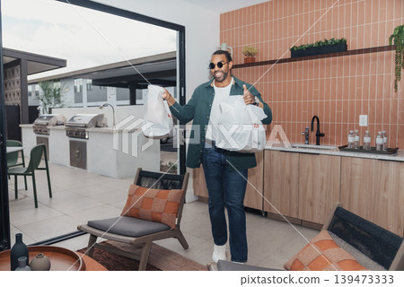 Man Arriving With Grocery Bags At Outdoor Kitchen, Wearing Sunglasses And Jacket, Smiling And Holding Multiple Paper Bags Modern Patio With Seating And Builtin Grill, Casual Weekend Vibe, 139473333