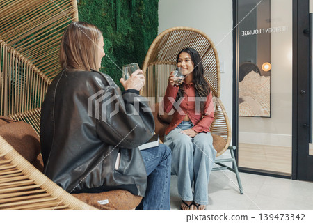 White And Asian Women Clinking Glasses For Brand Shoot, Content Creators Posing In Rattan Chairs With Editorial Styling, Leather Jacket And Blouse Outfits, Studio Backdrop With Poster, 139473342