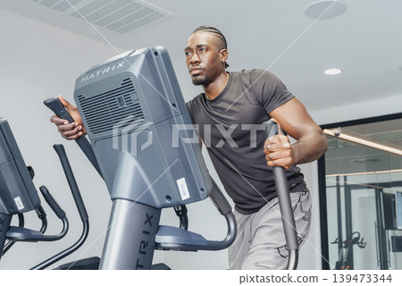 Black Man Checking Elliptical Console And Smartphone Metrics, Attentive Posture With One Hand On Handle And Eyes On Display, Modern Gym Interior, Focus On Datadriven Training And Progress Black Man Checking Elliptical Console And Smartphone Metrics, Attentive Posture With One Hand On Handle And Eyes On Display, Modern Gym Interior, Focus On Datadriven Training And Progress 139473344