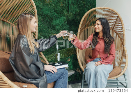 White And Asian Women Clinking Glasses In Rattan Chairs, Celebrating Creative Partnership, Startup Founders Exchanging Ideas, Leather Jacket And Pink Blouse Outfits, Notebook On Lap, Indoor White And Asian Women Clinking Glasses In Rattan Chairs, Celebrating Creative Partnership, Startup Founders Exchanging Ideas, Leather Jacket And Pink Blouse Outfits, Notebook On Lap, Indoor 139473364