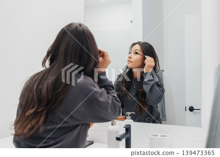 Casual Morning Mirror Application. Woman Applying Mascara In Gentle Light. Woman Carefully Applying Mascara During Morning Routine In Soft Natural Illumination With Sink Visible Casual Morning Mirror Application. Woman Applying Mascara In Gentle Light. Woman Carefully Applying Mascara During Morning Routine In Soft Natural Illumination With Sink Visible 139473365