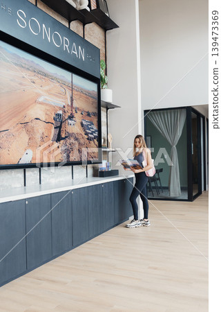 Woman Standing At Counter Reading Brochure, Modern Hotel Lobby With Oversized Sonoran Wall Art, Open Shelving With Plants And Decor, Light Wood Floor, Casual Jeans And Sneakers, Holding 139473369