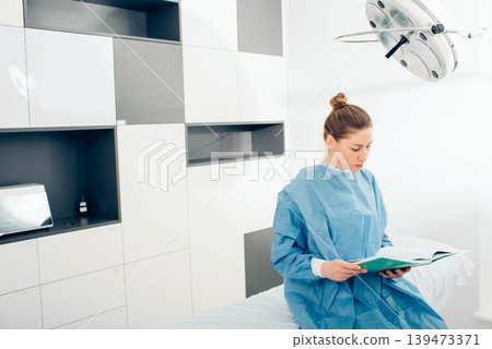 A medical professional in blue scrubs reads a patient file while seated on an examination table in a contemporary clinic A medical professional in blue scrubs reads a patient file while seated on an examination table in a contemporary clinic 139473371