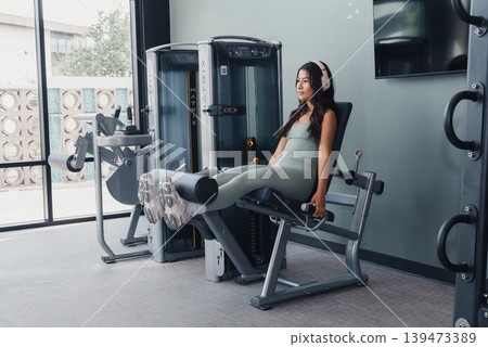 Woman Using Leg Extension Machine In Modern Gym, Focused Strength Set With Controlled Reps, Soft Natural Light, Fitted Activewear, Headphones, Clean Equipment And Bright Studio Atmosphere. 139473389