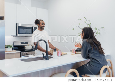 Asian Woman Animatedly Discussing Plans While Black Man Listens At Kitchen Island, Latte Cups And Jars On Counter, Bright Minimalist Space, Collaborative Planning And Everyday Strategy 139473404