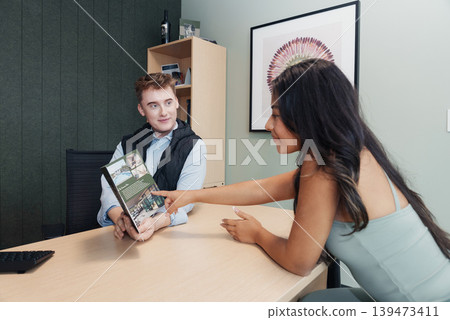 Client Pointing At Brochure In Meeting, Consultation Scene With Selection Of Property Images, Collaborative Decision Moment, Woman Indicating Preference And Discussing Options With Advisor. Client Pointing At Brochure In Meeting, Consultation Scene With Selection Of Property Images, Collaborative Decision Moment, Woman Indicating Preference And Discussing Options With Advisor. 139473411