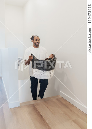 Man Carrying Filled Laundry Basket Indoors, Simple Home Setting With Soft Light, Casual Outfit And Content Expression Routine Chore Scene Showing Folded Linens, Tidy Staircase, Efficient Man Carrying Filled Laundry Basket Indoors, Simple Home Setting With Soft Light, Casual Outfit And Content Expression Routine Chore Scene Showing Folded Linens, Tidy Staircase, Efficient 139473418