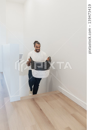 Man Carrying Laundry Basket Up Stairs, Bright Hallway, Hardwood Floors, Casual Tshirt, Thumbs Up, Welcoming Energy Woven Basket Filled With Linens, Natural Light Pouring From Landing, Man Carrying Laundry Basket Up Stairs, Bright Hallway, Hardwood Floors, Casual Tshirt, Thumbs Up, Welcoming Energy Woven Basket Filled With Linens, Natural Light Pouring From Landing, 139473419