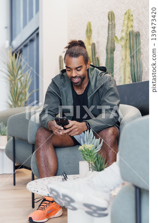 Man On Sofa Checking Phone Messages, Freelance Developer Managing Client Communication, Casual Hoodie And Sneakers, Focused Smile, Natural Light And Cactus Accents, Mobile Productivity 139473420