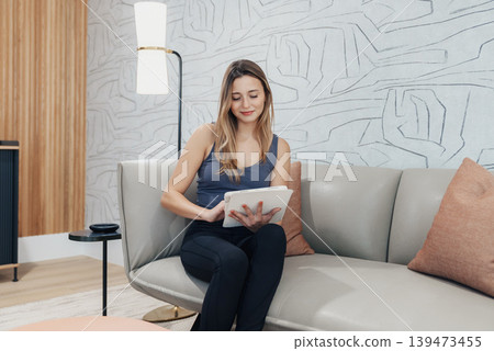 Woman On Sofa Reading Tablet, Content Creator Reviewing Portfolio And Captions, Soft Lamp Light, Relaxed Posture, Neutral Decor And Casual Outfit, Focused Browsing And Note Taking. 139473455