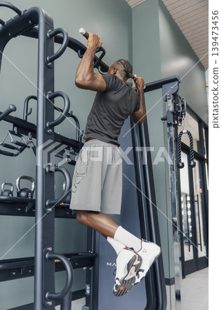 Black Man Doing Pullups On Bar With Fullrange Motion, Tense Grip And Focused Expression, Gym Rack And Weight Area In Background, Emphasis On Muscle Engagement, Power And Disciplined Strength Black Man Doing Pullups On Bar With Fullrange Motion, Tense Grip And Focused Expression, Gym Rack And Weight Area In Background, Emphasis On Muscle Engagement, Power And Disciplined Strength 139473456